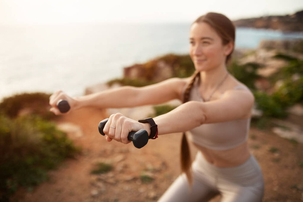Positive energy young caucasian lady in sportswear doing arm exercise with dumbbells, enjoy training on ocean beach, motivating. Body care, health care, fitness and workout outdoor - Photo, image