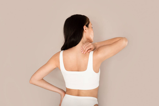 Tired young woman in white comfortable underwear massaging shoulder, have pain in upper back, isolated on beige studio background, copy space, back view. Poor posture, muscle overuse - Foto, Imagen