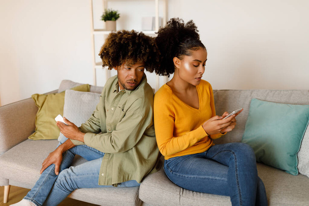 Jealousy Problem. African American Boyfriend Peeking At Girlfriends Phone Reading Messages Suspecting Infidelity Sitting On Couch At Home. Jealous Man Spying On Cheating Woman - Foto, imagen