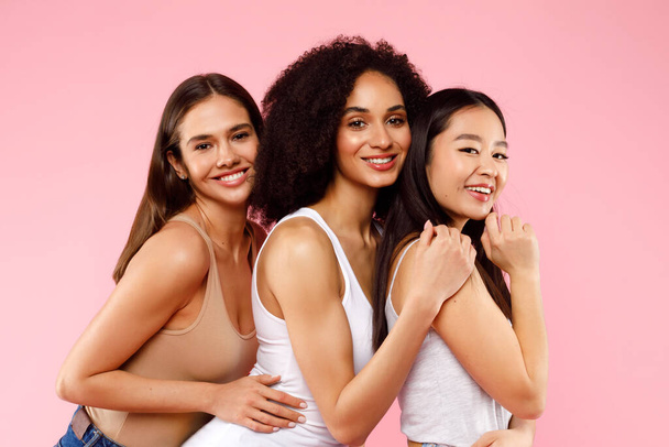 Счастливые три разные дамы позируют и обнимаются, стоя на розовом студийном фоне и улыбаясь на камеру. Концепция женской дружбы - Фото, изображение