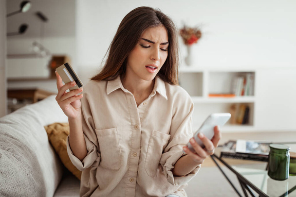 Upset sad millennial european female with credit card looking at phone in minimalist living room interior. Problems with online shopping, finance banking, mistake in order, sale - 写真・画像