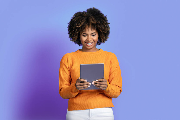 Smiling beautiful young african american lady with bushy hair wearing stylish casual outfit using modern pad digital tablet on purple studio background, reading blog on Internet, copy space - Foto, Imagem