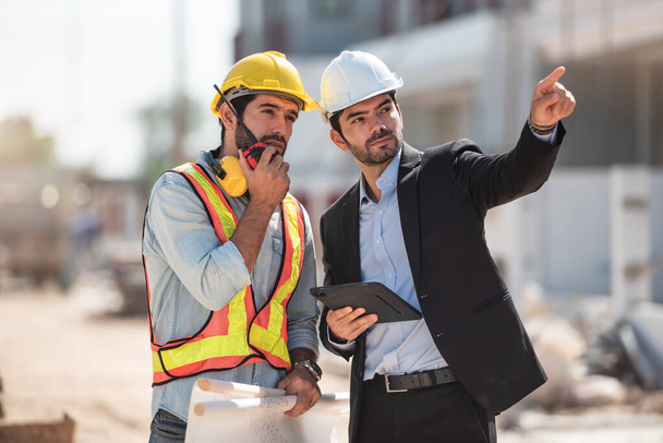 Mérnök és építész csapat együtt dolgozik az építkezésen, két fiatal munkavezető sisakot visel, és az ingatlaniparról beszélgetnek.. - Fotó, kép