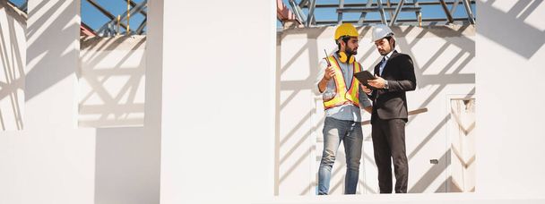 Hombres de trabajo en equipo en el sitio de construcción, dos ingenieros civiles en casco de seguridad casco duro usando tableta digital y plano de trabajo mientras están de pie en la fábrica. - Foto, imagen