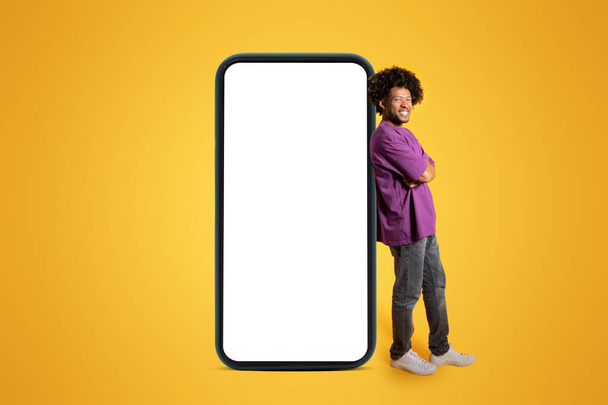 Cheerful confident adult african american curly man in purple t-shirt stands near huge smartphone with blank screen isolated on yellow studio background. Ad and offer, app, blog, website and sale - Valokuva, kuva