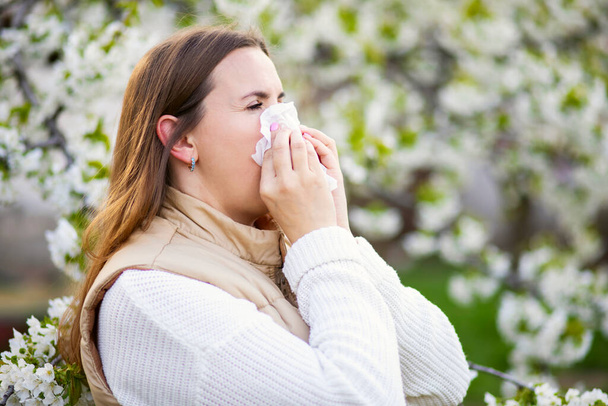 Kichająca kobieta cierpiąca na sezonową alergię wiosną z wycieraczką do nosa wśród kwitnących drzew w parku. Pojęcie alergii wiosennej - Zdjęcie, obraz