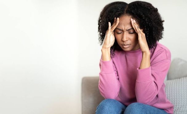 Sad tired young african american curly woman sitting on sofa, presses hands to temples, suffering from headache in living room interior, copy space. Health problems, stress, migraine at home - Photo, Image