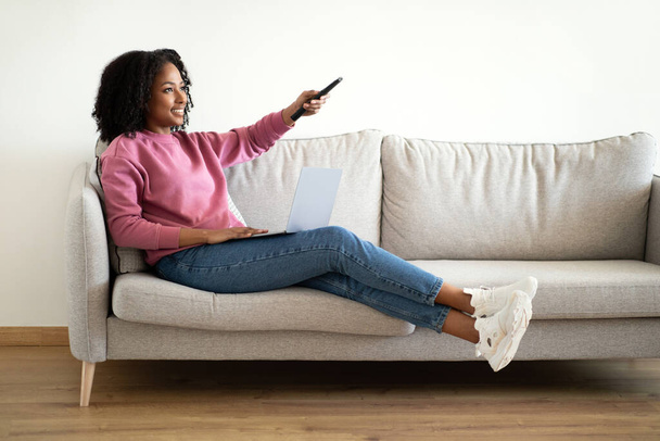 Улыбающаяся молодая африканская кудрявая женщина, сидящая на диване с пультом, наслаждается свободным временем и телешоу в минималистичном интерьере гостиной, свободным пространством. Расслабьтесь, отдыхайте дома, вечер кино - Фото, изображение