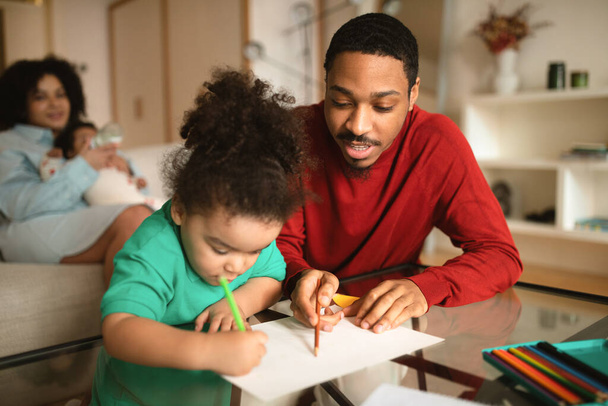 Boldog afro-amerikai család időt töltenek együtt otthon, jóképű Y-generációs fekete férfi rajzol vagy színez a kisfiával, anya pihen a kanapén, eteti a csecsemőt, másolja a helyet - Fotó, kép