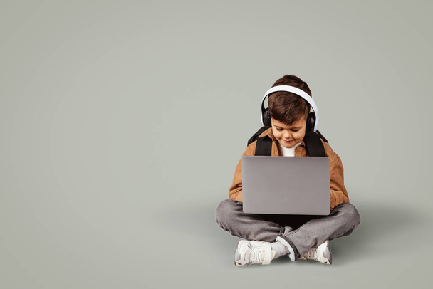 6 yaşında, kablosuz kulaklıklarıyla çantasıyla dizüstü bilgisayarda yazan, internetten oyun oynayan, gri arka planda izole edilmiş, Avrupalı bir çocuk. Ders, bilgi, eğitim. - Fotoğraf, Görsel