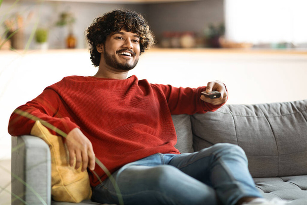 Weekend Pastime. Cheerful Indian Guy Sitting On Couch And Watching Tv, Smiling Young Eastern Man Holding Remote Controller, Switching Channels While Resting On Sofa At Home, Free Space - Foto, imagen