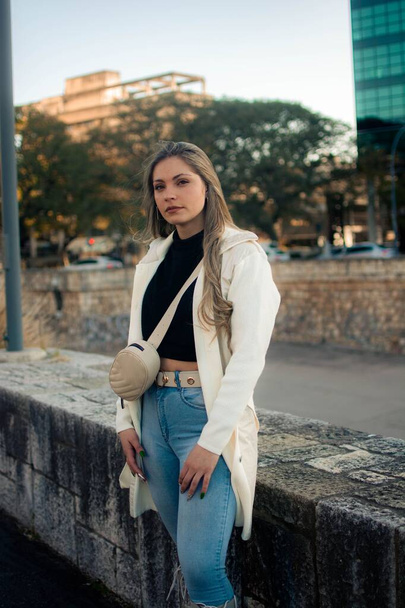 A vertical of a beautiful Argentinian woman in a white coat and blue jeans outdoors - Фото, зображення