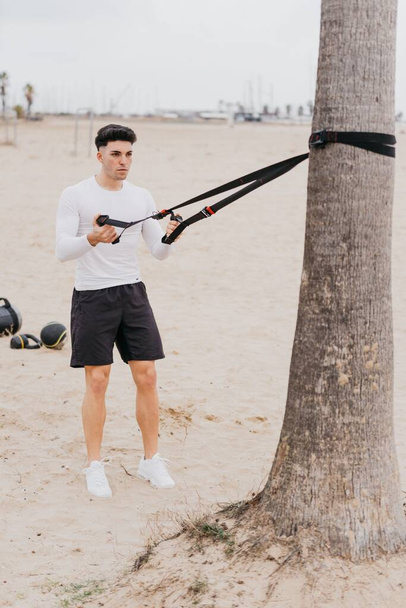 Nuori istuvuus espanjalainen urheilullinen mies tekee olkapään vedä harjoituksia köydellä rannalla - Valokuva, kuva