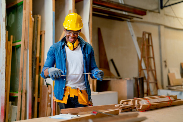 Afroamerykanin stolarz używa miernika taśmowego do pracy w fabryce ze szczęściem, uśmiechając się podczas pracy. - Zdjęcie, obraz