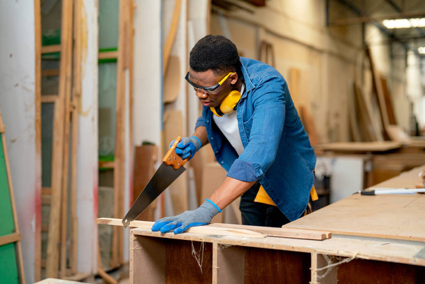 Breed beeld van Afro-Amerikaanse man als timmerman gebruik zaag om hout te snijden in de fabriek of op het werk met geluk en uitdrukkelijke glimlach tijdens het werk. - Foto, afbeelding