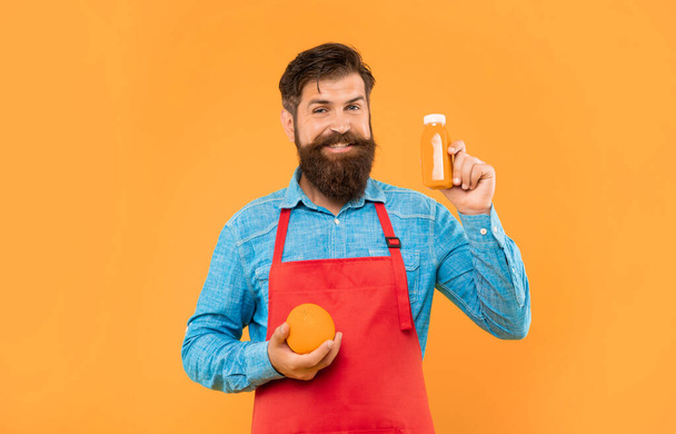 Happy man in apron holding orange and juice bottle yellow background, bartender. - 写真・画像