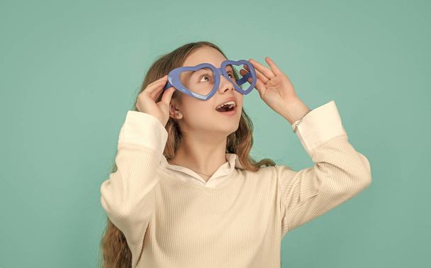 Surprised child looking through big funny heart-shaped glasses blue background. - Zdjęcie, obraz