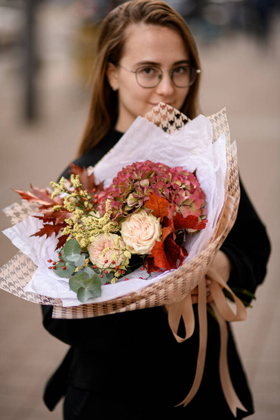 Young woman holding big and beautiful autumn style bouquet of fresh white roses, hydrangea , dog-rose, gypsophila and oak leaves deorater with white abd brown wrapping paper with light riband - 写真・画像