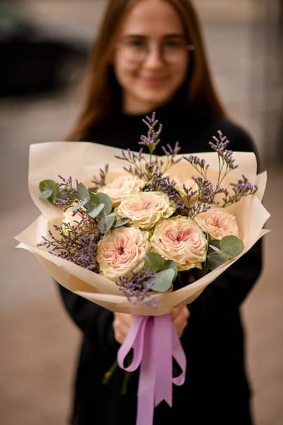Mujer joven muy agradable vestida de negro sosteniendo gran ramo de flores de rosas cremosas frescas, limonio y eucalipto envuelto en papel ligero decorado con cinta violeta - Foto, imagen
