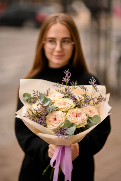 Young woman dressed in black holding big flower bouquet of fresh creamy roses, limonium and eucaliptus on street background - Zdjęcie, obraz