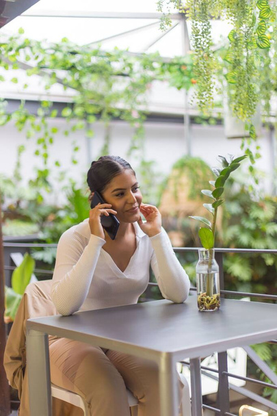 A vertical shot of a young Hispanic woman sitting near the table and talking to the phone in garden - Foto, afbeelding
