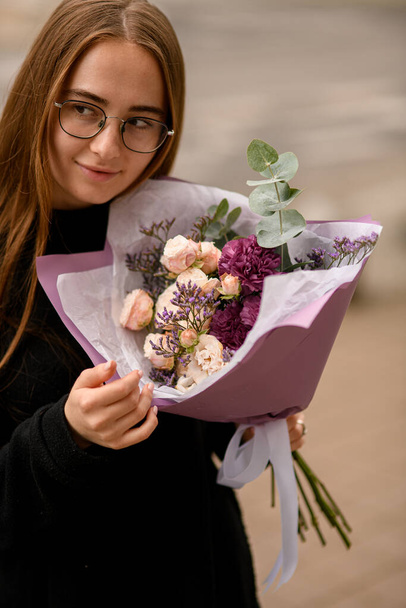 Nice girl gently holding big and beautiful bouquet of fresh roses, eucaliptus, limonium and carnations in cream, violet, and burgundy colors - Zdjęcie, obraz