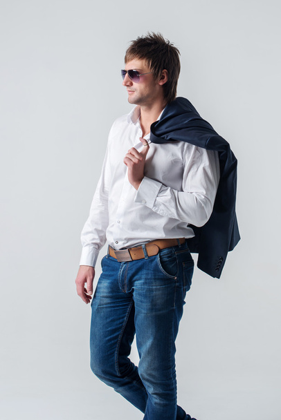 Σοβαρή αυτοπεποίθηση ωραίος τύπος ελκυστική στο τζιν κρατώντας κοστούμι σακάκι στον ώμο του αναζητούν μακριά αριστερή πλευρά μέσα από γυαλιά ηλίου στέκεται ενάντια σε τοίχο - Φωτογραφία, εικόνα