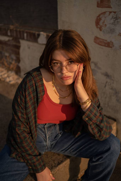 A vertical closeup of an attractive female with glasses sitting on the sidewalk blurred background - 写真・画像