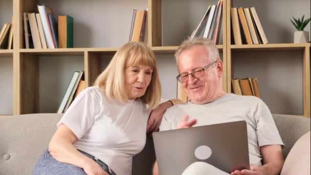 Ευτυχισμένο ζευγάρι ηλικιωμένων που κάθονται στο σαλόνι και κοιτάζουν το λάπτοπ. Η παλαιότερη γενιά και τεχνολογία. Αναζήτηση στο Διαδίκτυο. - Πλάνα, βίντεο