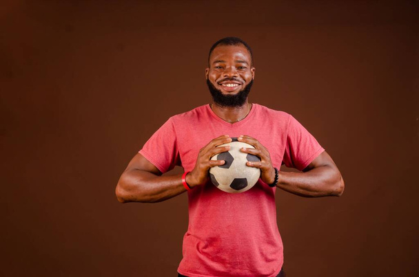 An attractive happy African man holding a soccer ball isolated on a brown background. - Foto, Bild