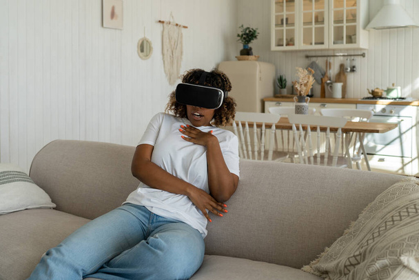 Nadšený divil africká americká žena virtuální brýle sledování filmu hraje videohry. Usmívající se žena se dívá na virtuální svět ve VR helmě sedí na gauči doma. Cyberspace, koncept zábavy. - Fotografie, Obrázek