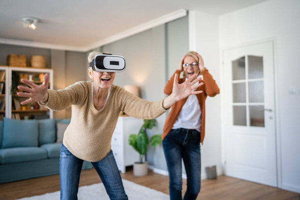 дві жінки зрілі старші кавказькі жінки друзі або сестри вдома насолоджуються гарнітурою віртуальної реальності VR реальні люди активні старші мають веселу концепцію дозвілля
 - Фото, зображення