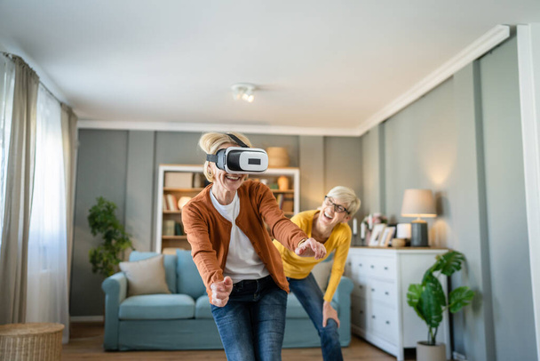 dos mujeres maduras mayores caucásicas amigas o hermanas en casa disfrutar de realidad virtual VR auriculares personas reales activos senior divertirse concepto de ocio - Foto, imagen