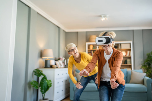 dos mujeres maduras mayores caucásicas amigas o hermanas en casa disfrutar de realidad virtual VR auriculares personas reales activos senior divertirse concepto de ocio - Foto, Imagen