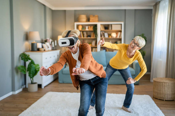 two women mature senior caucasian female friends or sisters at home enjoy virtual reality VR headset real people active senior having fun leisure concept - Fotoğraf, Görsel