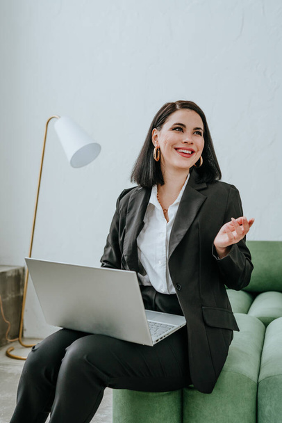 Lächelnde Geschäftsfrau im Anzug mit Laptop sitzt auf einem grünen Sofa. Im Hintergrund eine weiße Wand und eine weiße Stehlampe. - Foto, Bild