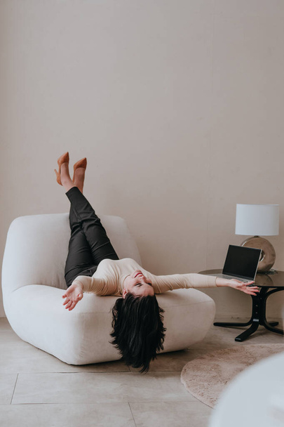 Young smiling brunette woman in business clothes lying on an armchair with feet up and spreads her arms indoor. Selective focus. - Фото, зображення