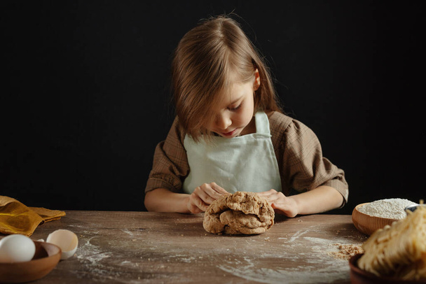 Little girl wearing apron kneading dough on wooden background against dark wall. Child prepairing homemade bakery or noodles. Selective focus. - Фото, зображення