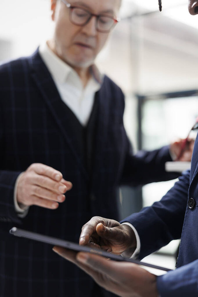 Senior man asking employee to check inventory for shirt in shopping centre, client wanting to buy new collection. African american showroom manager analyzing clothes parcel on tablet. Close up - 写真・画像