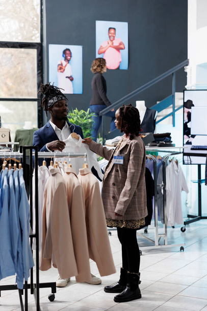 Boutique worker ayuda a los clientes con estilo con la elección de la camisa blanca durante la actividad jalá. Cliente afroamericano que compra ropa y accesorios de moda en el centro comercial. Concepto de moda - Foto, imagen