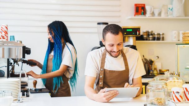 Smiling man in apron using tablet while accepting online order in cafe. African American female barista making coffee on the background in coffee shop - Foto, Imagem