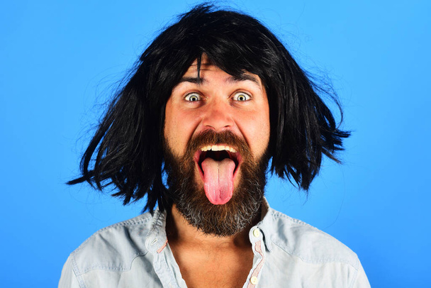 Zabawny brodaty mężczyzna w czarnej peruce pokazuje język. Szczęśliwy człowiek z brodą i wąsami w peruce. Emocje ludzi. Twarzą do twarzy. Uczucia i emocje. Hipster w czarnym periwigu. Stylowy hipster z czarnymi włosami - Zdjęcie, obraz