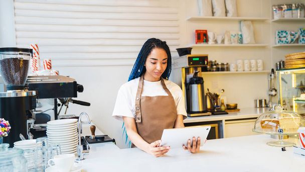 Pretty young female barista standing at the counter while holding tablet and looking at the screen. Online order concept - Photo, Image
