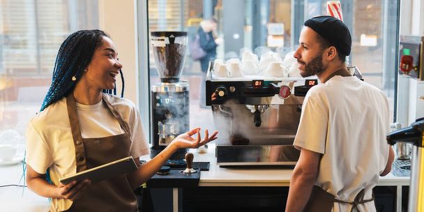 Happy young woman in apron using digital tablet while talking with male barista in coffee shop. Online order concept - Foto, afbeelding
