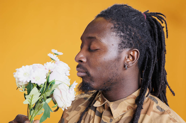 Υπέροχος άνθρωπος κρατώντας λουλούδια μπουκέτο μυρίζοντας τριαντάφυλλα, ενώ θέτουν σε στούντιο πάνω από κίτρινο φόντο. Αυθεντικός Αφροαμερικάνος που γιορτάζει την ημέρα του Αγίου Βαλεντίνου, απολαμβάνοντας την αγάπη του. - Φωτογραφία, εικόνα