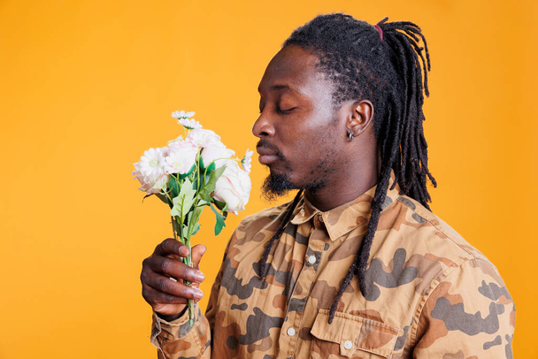 African american man smelling white roses bouquet, posing with flowes for valentine s day in studio over yellow background. Smiling romantic young adult enjoying love holiday - Zdjęcie, obraz