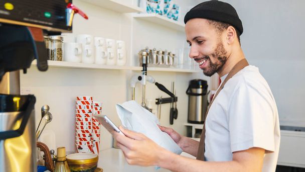 Young smiling barista holding craft package with coffee beans in coffee shop. Man looking at tablet screen while making online order - Valokuva, kuva