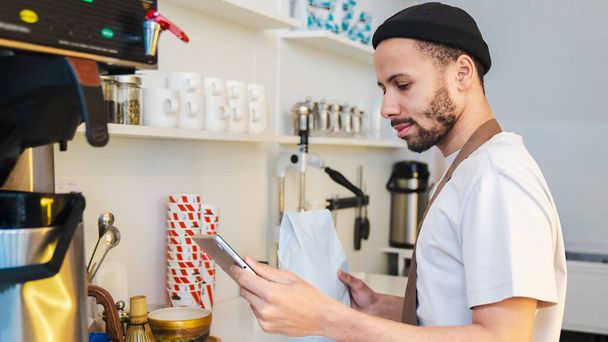 Side view of male barista using digital tablet while taking online order and holding package with coffee beans. Business restaurant food and drink concept - Fotó, kép