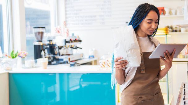 Smiling female barista making online order while working at coffee shop. Lady holding paper package with coffee beans. Business restaurant food and drink concept - Photo, Image