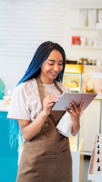 Smiling female barista holding paper package with coffee beans while using tablet, making online order in coffee shop - Foto, Imagen